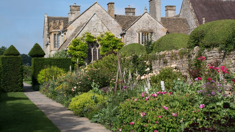 Part of the garden at Lytes Cary Manor, with a path past an old stone wall, flowerbeds, decorative hedges and the manor in the background, Somerset
